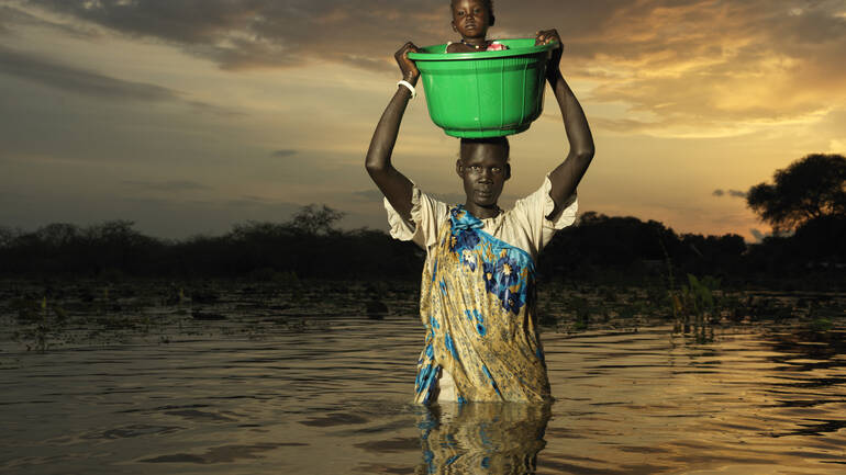 Eine Frau aus dem Südsudan steht in den Fluten und trägt ihr kleines Kind in einer grünen Wanne über dem Kopf