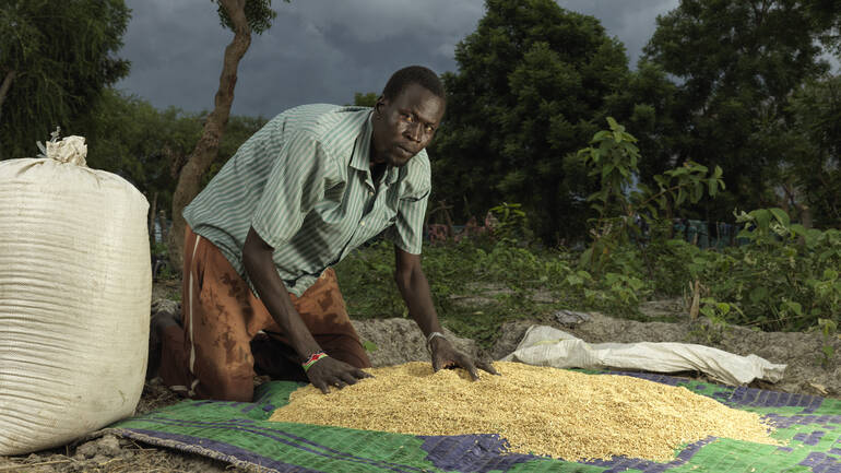 James Wuor aus Paguir im Südsudan kniet auf einer Decke, auf der er Reissamen sortiert. Er schaut in die Kamera.