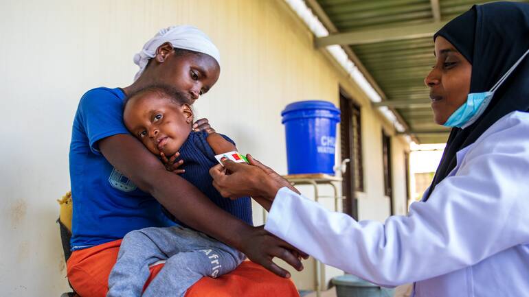 Khadija, ein Mädchen aus Kenia, wird auf dem Schoß ihrer Mutter sitzend von einer Krankenschwester mit einem MUAC-Band untersucht.