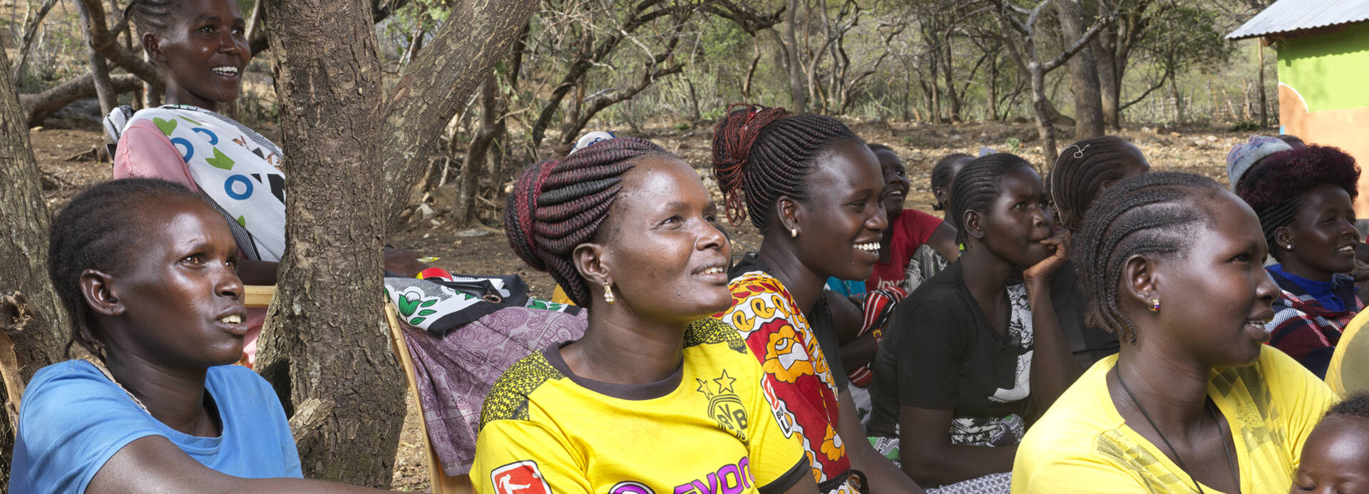 Mehrere Frauen aus Kenia sitzen in ihrem Dorf und hören offenbar jemandem zu, ein Lächeln auf ihren Lippen.