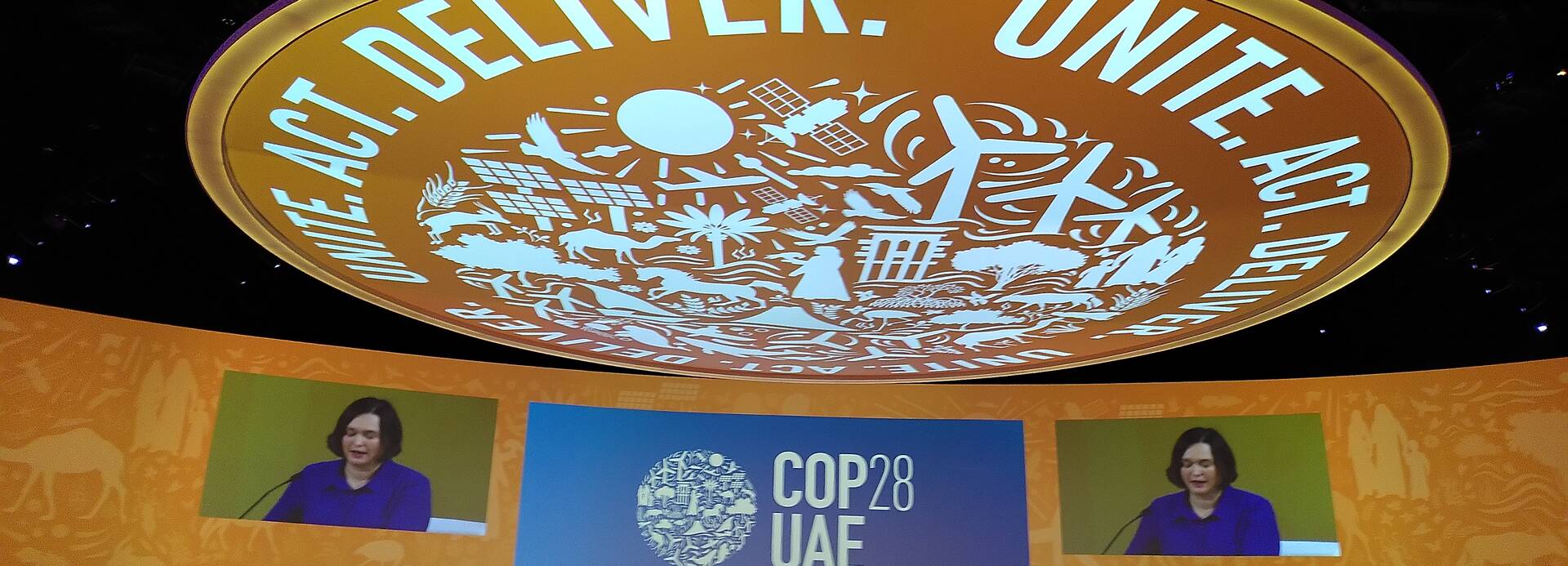 Im Plenarsaal der Klimakonferenz in Dubai sitzen die Vertreter*innen der Staaten und hören der Sprecherin vorne zu. Über allen wölbt sich eine große Kuppel mit der Aufschrift "Unite, Act, Deliver".