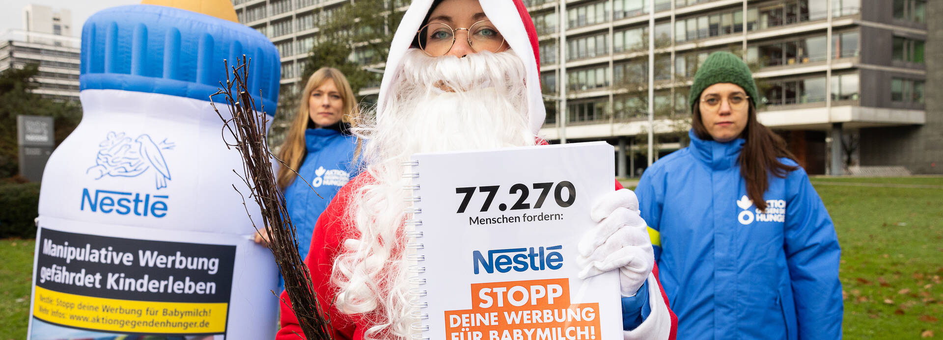 Der Nikolaus hält ein Buch mit den 77.270 Unterschriften gegen Nestlés Werbestrategie hoch, im Hintergrund eine große Babyflasche und zwei Mitarbeiterinnen von Aktion gegen den Hunger - alle vor dem Nestlé-Hauptsitz in Frankfurt am Main.