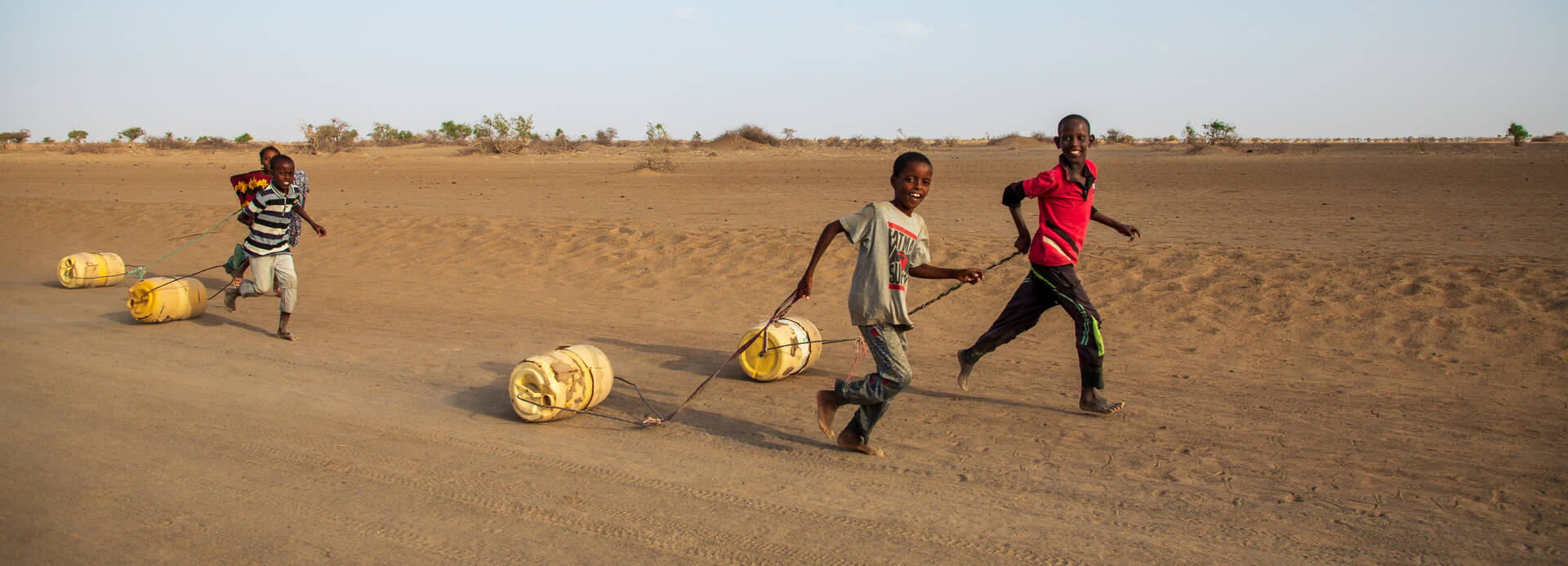 Vier Jungs aus Kenia laufen mit Wassertanks an Seilen über einen Wüstenweg.