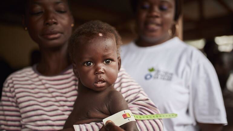 Kind mit Muac im Arm der Mutter und Mitarbeiterin von Aktion gegen den Hunger