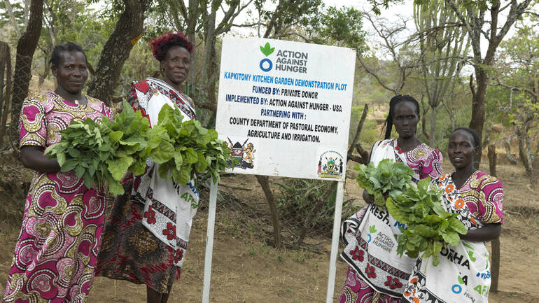 Vier Frauen aus dem Dorf Kapkitony in Kenia stehen mit Gemüse in der Hand neben einem Schild von Aktion gegen den Hunger.