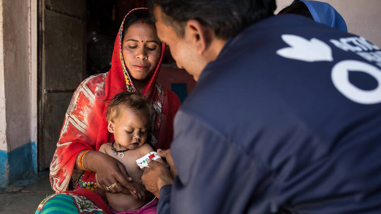 Bhumika sitzt auf dem Schoß ihrer Mutter, während ein Gesundheitshelfer von Aktion gegen den Hunger ihren Arm mit einem Muac-Band vermisst – das Band zeigt den roten Bereich an: Mangelernährung.