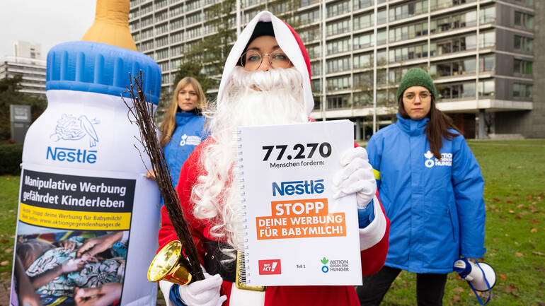 Der Nikolaus hält ein Buch mit den 77.270 Unterschriften gegen Nestlés Werbestrategie hoch, im Hintergrund eine große Babyflasche und zwei Mitarbeiterinnen von Aktion gegen den Hunger - alle vor dem Nestlé-Hauptsitz in Frankfurt am Main.