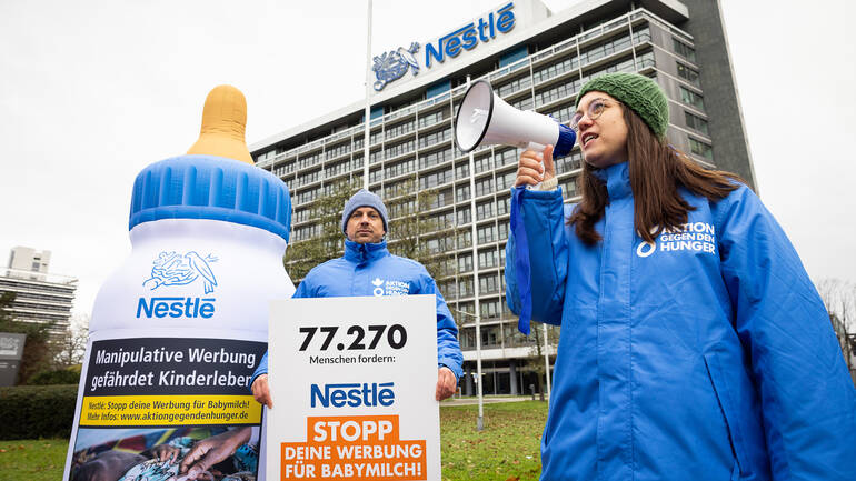 Eine Mitarbeiterin von Aktion gegen den Hunger ruft in ein Megafon, während ein Mitarbeiter das Kampagnenziel auf einem Schild hochhält, daneben eine aufblasbare Babymilch-Flasche.