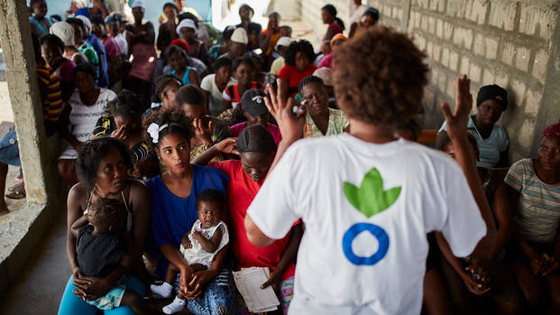 Eine Mitarbeiterin von Aktion gegen den Hunger erklärt einer Gruppe Frauen, Männer und Kinder aus Haiti, wie sie sich mit gesunder Ernährung und Hygiene vor Cholera schützen können.