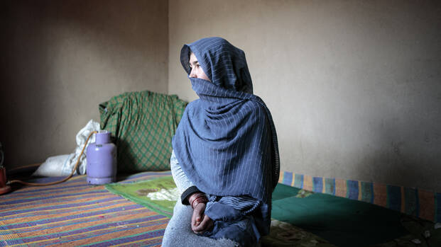 Eine Frau aus Afghanistan sitzt in ihrem Zuhause und schaut zur Seite. Ein blaues Tuch bedeckt ihren Kopf und ihren Mund.