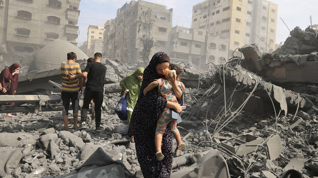 Eine Frau trägt ihr Kind durch eine von Angriffen zerstörte Straße in Gaza Stadt, im Hintergrund weitere Menschen.