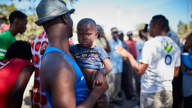 Ein Mann mit Hut trägt ein Kind auf dem Arm, während er einer Erklärung eines unserer Mitarbeiter in Haiti zuhört. 