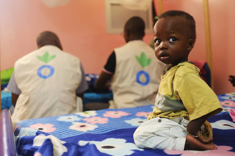 kind-sitzt-in-einer-station-von-aktion-gegen-den-hunger-auf-einer-liege-im-hintergrund-zwei-helfer kind-sitzt-in-einer-station-von-aktion-gegen-den-hunger-auf-einer-liege-im-hintergrund-zwei-helfer