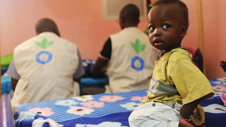 kind-sitzt-in-einer-station-von-aktion-gegen-den-hunger-auf-einer-liege-im-hintergrund-zwei-helfer kind-sitzt-in-einer-station-von-aktion-gegen-den-hunger-auf-einer-liege-im-hintergrund-zwei-helfer