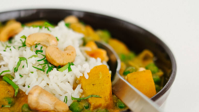 Eine Schüssel mit Süßkartoffel-Curry mit Erbsen, angerichtet mit Reis, Cashews und Kräutern, ein Löffel steckt im Essen.