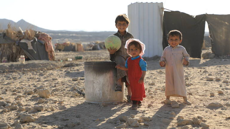 Drei Kinder mit einem Ball stehen auf einem Geröllfeld in einem Flüchtlingscamp im Jemen.