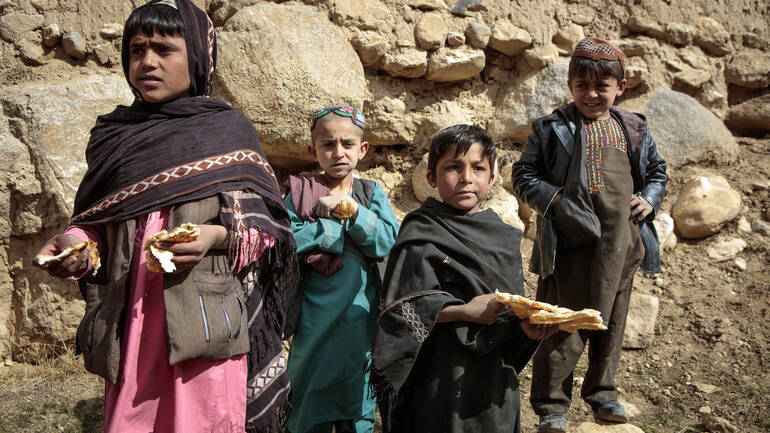 Vier Kinder aus Afghanistan stehen mit Brot in der Hand auf Steinen.