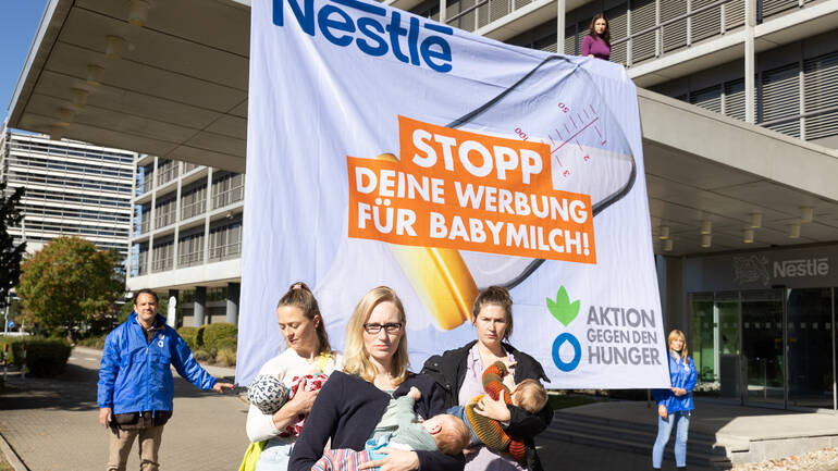 Nestlé: Stopp deine Werbung für Babymilch! Aktion gegen den Hunger mit Müttern und Babys vor der Nestlé-Zentrale Mütter mit Babys stillen vor der Nestlé-Zentrale in Frankfurt am Main, im Hintergrund halten Aktivist*innen von Aktion gegen den Hunger ein Banner: Nestlé: Stopp deine Werbung für Babymilch!