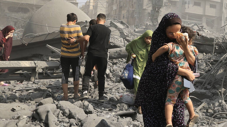 Eine Frau trägt ihr Kind durch eine von Angriffen zerstörte Straße in Gaza Stadt, im Hintergrund weitere Menschen.