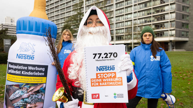 Der Nikolaus hält ein Buch mit den 77.270 Unterschriften gegen Nestlés Werbestrategie hoch, im Hintergrund eine große Babyflasche und zwei Mitarbeiterinnen von Aktion gegen den Hunger - alle vor dem Nestlé-Hauptsitz in Frankfurt am Main.