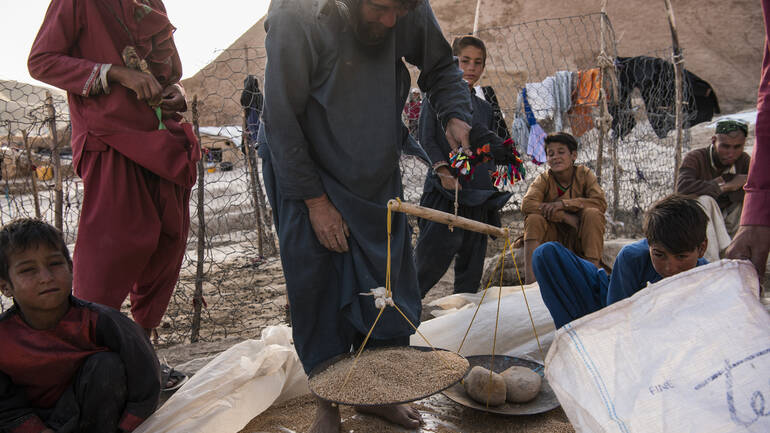Ein Mann wägt Weizen mithilfe einer Waage ab, rundherum sitzen Kinder.