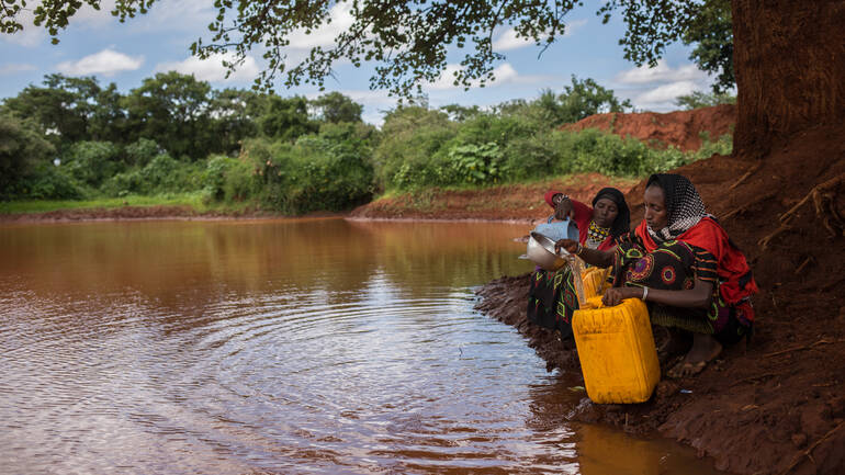 Zwei Frauen schöpfen sichtbar verschmutztes Wasser aus einem See