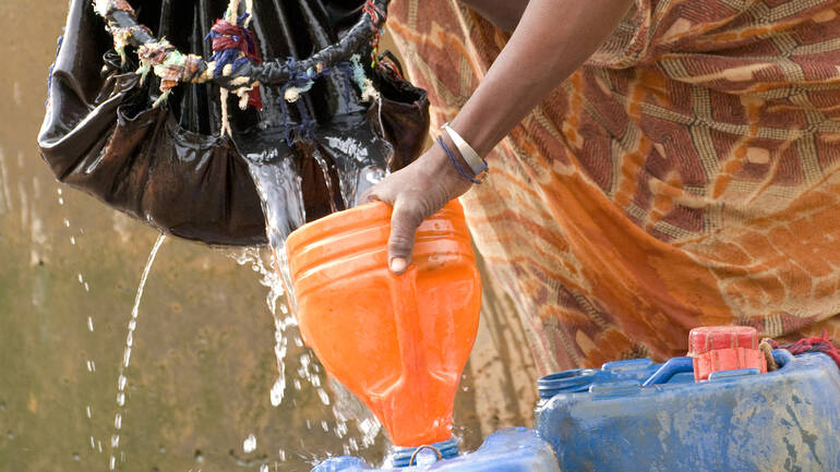 Wasser filtern hilft gegen grobe Verschmutzungen. Zwei Frauen filtern Brunnenwasser durch einen Filter und gießen es mithilfe eines Trichters in einen Kanister.