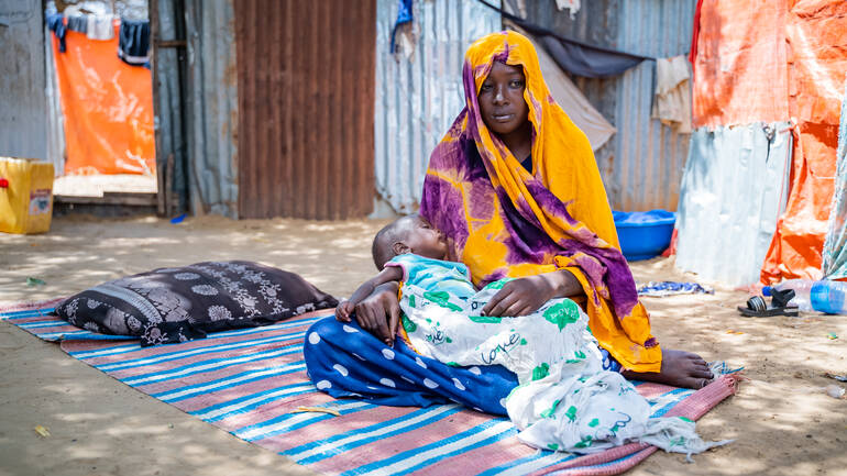Eine somalische Mutter sitzt in buntes Tuch gewickelt mit ihrem Kind im Schoß auf einem Teppich in einer Unterkunft aus Wellblech