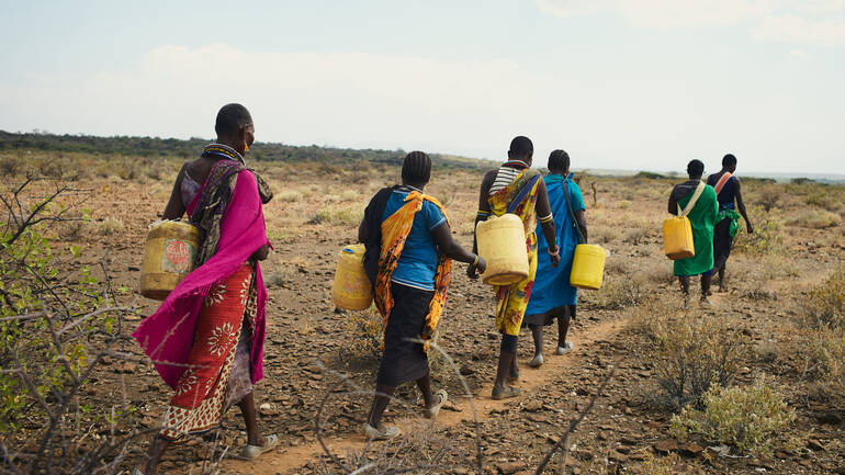 Sechs Frauen aus Kenia sind mit Kanistern auf dem Weg durch eine karge Landschaft, um Wasser an einem Wasserloch zu holen.