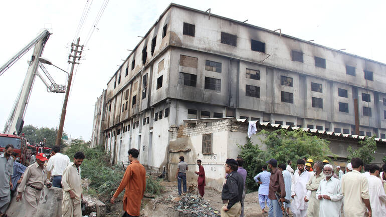 Die Fabrik Ali Enterprises in Pakistan brennt, Arbeiter und Feuerwer stehen davor.