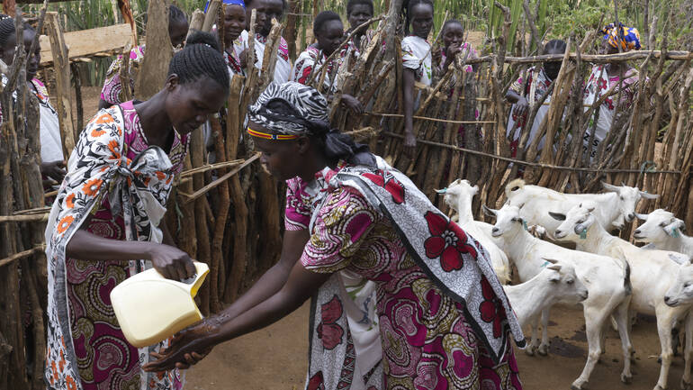 Eine Mutter aus dem Dorf hilft Rosalyne beim Händewaschen, im Hintergrund Ziegen und hinter dem Zaun Mütter und Kinder aus dem Dorf.