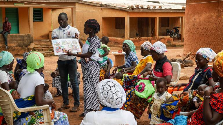 Eine Frau von der Côte d'Ivoire berät andere Frauen zum Stillen. Eine Frau mit Baby auf dem Rücken berät andere Frauen mit Kindern zum Stillen. Alle sitzen um sie herum, im Hintergrund ein Mann.