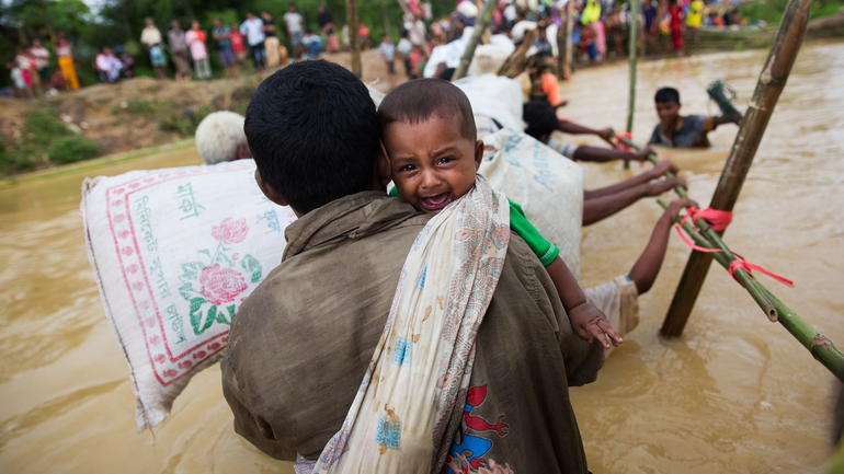 Ein Vater trägt sein weinendes Kind und einen Sack durch ein nach Zyklon Mocha überflutetes Camp.