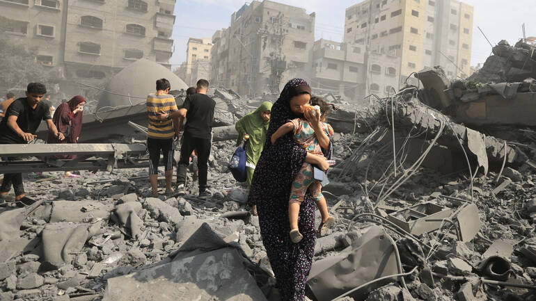 Eine Frau trägt ihr Kind durch eine von Angriffen zerstörte Straße in Gaza Stadt. Eine Frau trägt ihr Kind durch eine von Angriffen zerstörte Straße in Gaza Stadt, im Hintergrund weitere Menschen.
