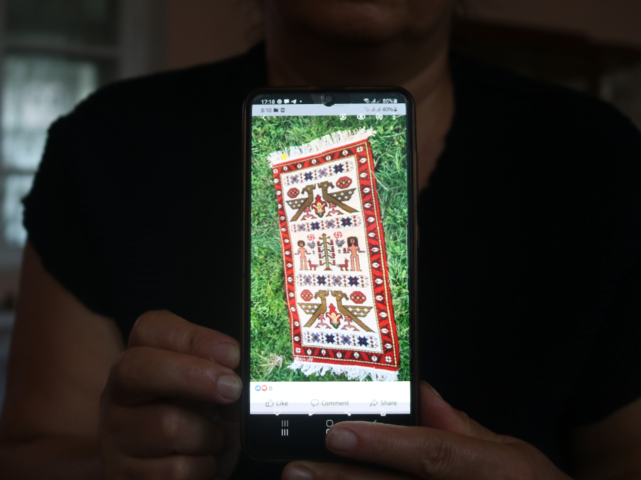 Nazik zeigt einen der Teppiche, den sie gewebt hat. Eine Person hält den Bildschirm eines Smartphones in die Kamera, auf dem ein handgewebter Teppich zu sehen ist.
