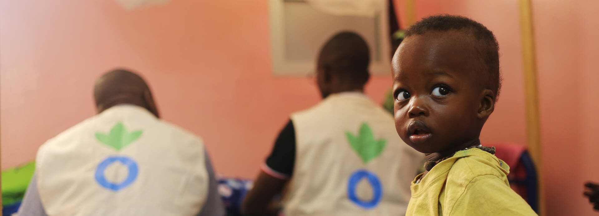Aktion gegen den Hunger unterstützt die Menschen vor Ort – wie diesen kleinen Jungen aus dem Senegal. Ein kleiner Junge sitzt auf einer geblümten Decke, im Hintergrund zwei Mitarbeiter von Aktion gegen den Hunger