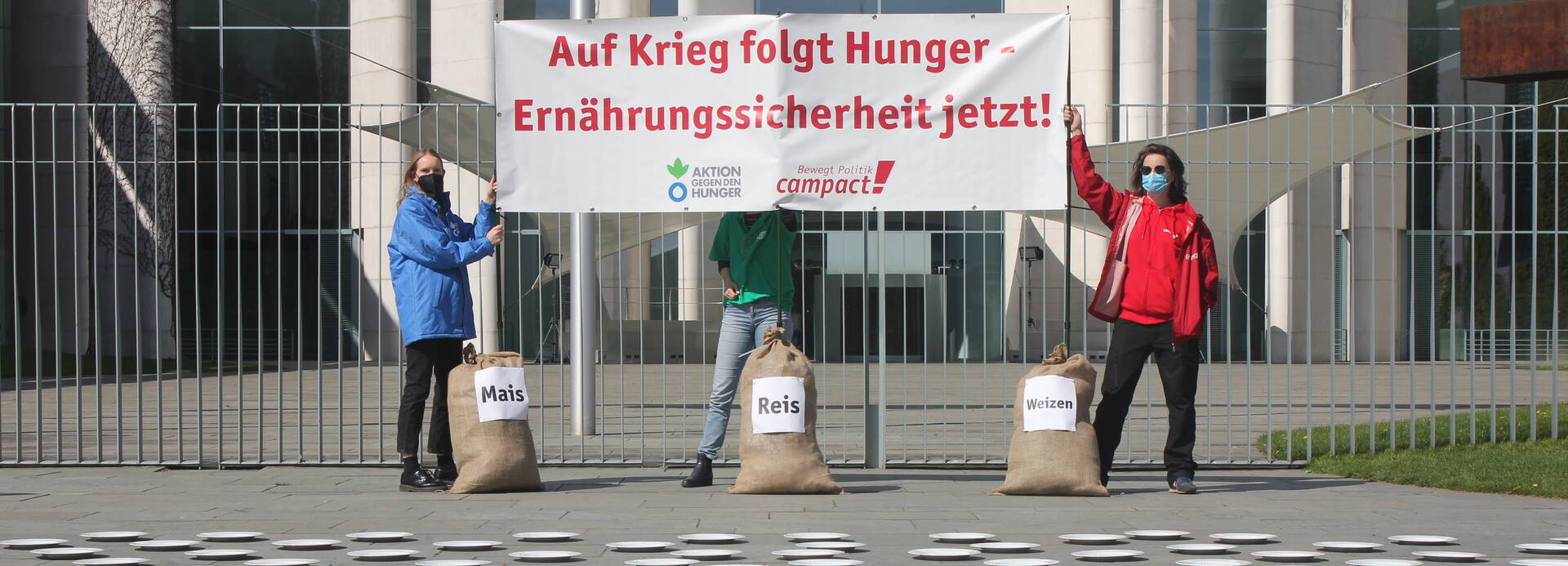 Drei Aktivist*innen von Aktion gegen den Hunger und Campact mit Banner "Auf Krieg folgt Hunger – Ernährungssicherheit jetzt!", mit drei Säcken Mais, Reis und Weizen vor 282 Tellern.