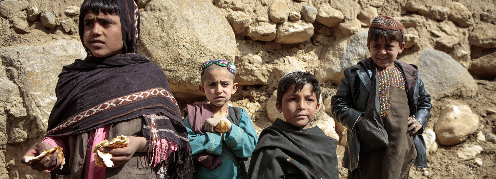 Vier Kinder aus Afghanistan stehen mit Brot in der Hand auf Steinen.
