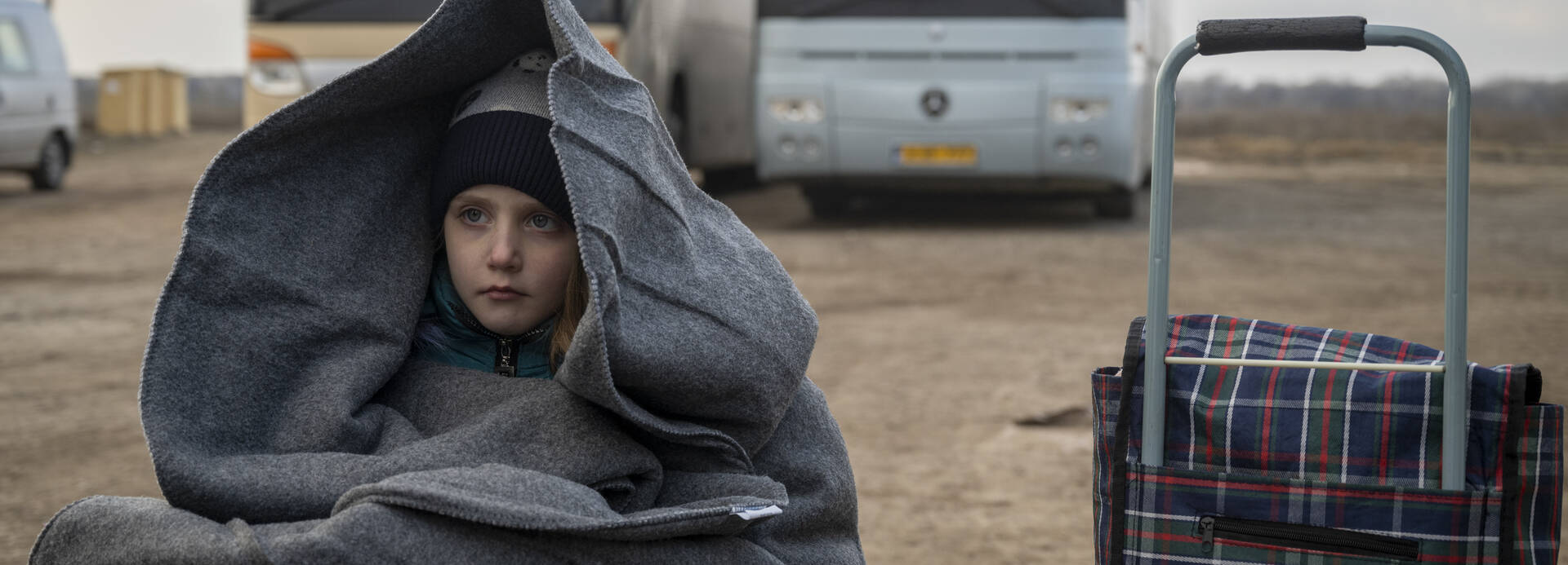 Ein Mädchen sitzt in eine Decke gehüllt neben einem Rollkoffer vor zwei Bussen an der Grenze während der Flucht aus der Ukraine.