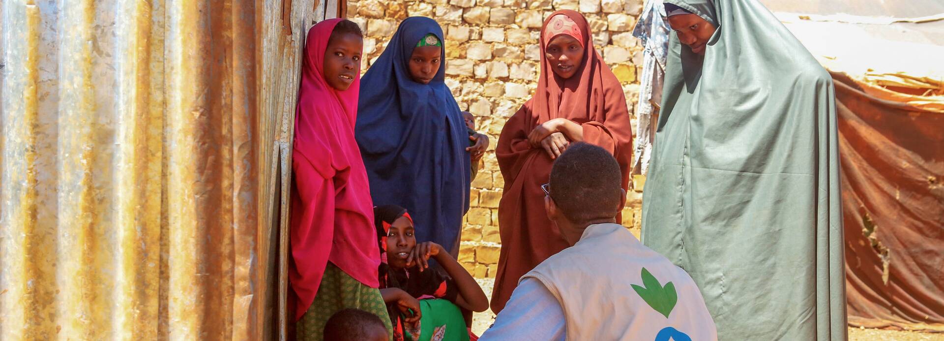 Ein Mitarbeiter von Aktion gegen den Hunger ist im Gespräch mit Frauen und Kindern aus Somalia – alle suchen Schatten hinter einer Wellblechwand.