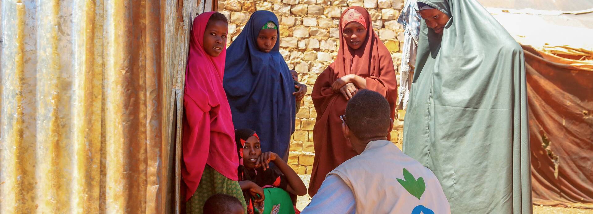 Die Hilfsorganisation Aktion gegen den Hunger ist seit 1992 in Somalia im Einsatz, um Mangelernährung zu diagnostizieren und zu behandeln. Hier berät eine Gesundheitsfachkraft Mütter, die durch Dürre und Konflikt aus ihrem Zuhause vertrieben wurden. Ein Mitarbeiter von Aktion gegen den Hunger ist im Gespräch mit Frauen und Kindern aus Somalia – alle suchen Schatten hinter einer Wellblechwand.