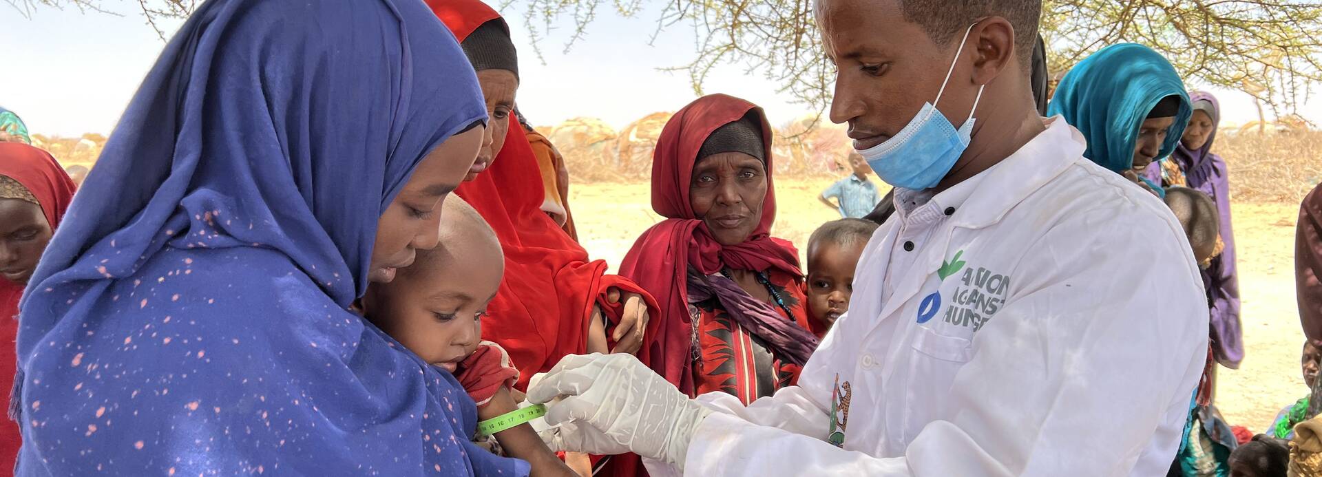 Mütter aus Somalia lassen ihre Kinder von Aktion gegen den Hunger auf Mangelernährung untersuchen. Ein Mitarbeiter von Aktion gegen den Hunger untersucht in Somalia ein Kleinkind auf den Armen seiner Mutter auf Mangelernährung, im Hintergrund warten weitere Mütter mit Kindern.