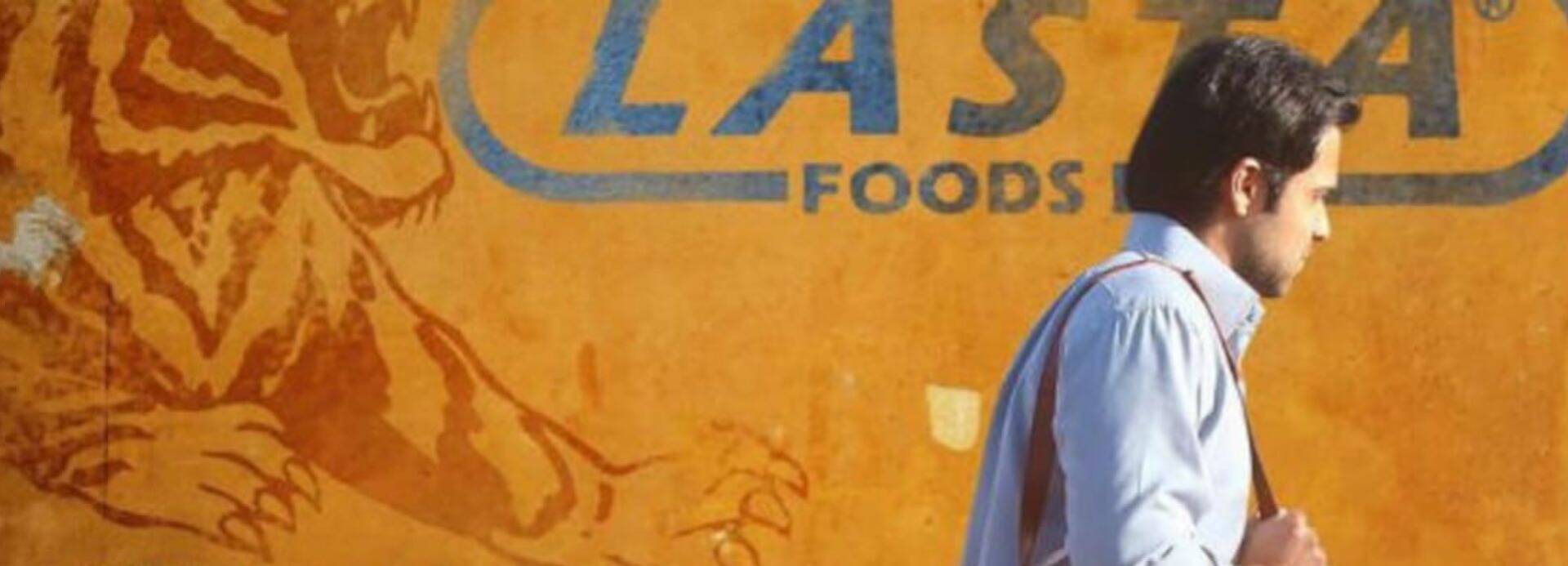 Ayan vor dem Logo seines Arbeitgebers, des Lebensmittelkonzerns Lasta Ayan, Protagonist aus dem Film Tigers, geht mit gesenktem Kopf am Logo des Lebensmittelkonzerns Lasta vorbei