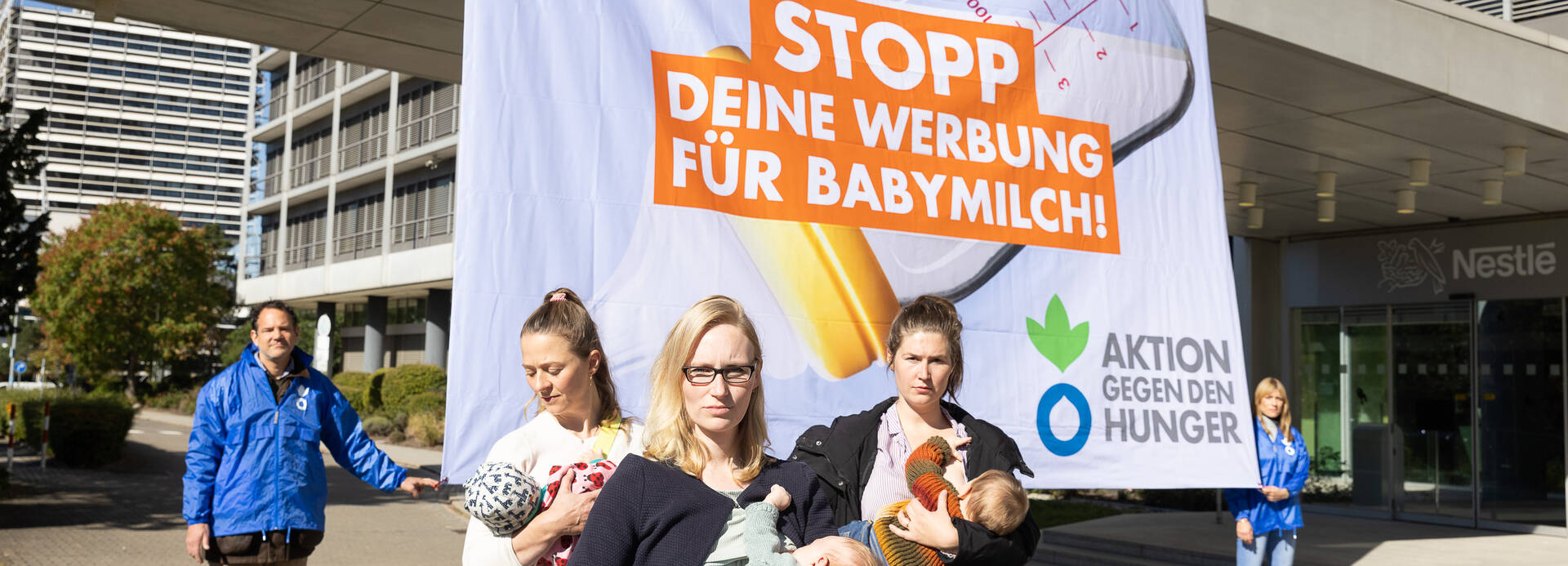 Mütter mit Babys stillen vor der Nestlé-Zentrale in Frankfurt am Main, im Hintergrund halten Aktivist*innen von Aktion gegen den Hunger ein Banner: Nestlé: Stopp deine Werbung für Babymilch!