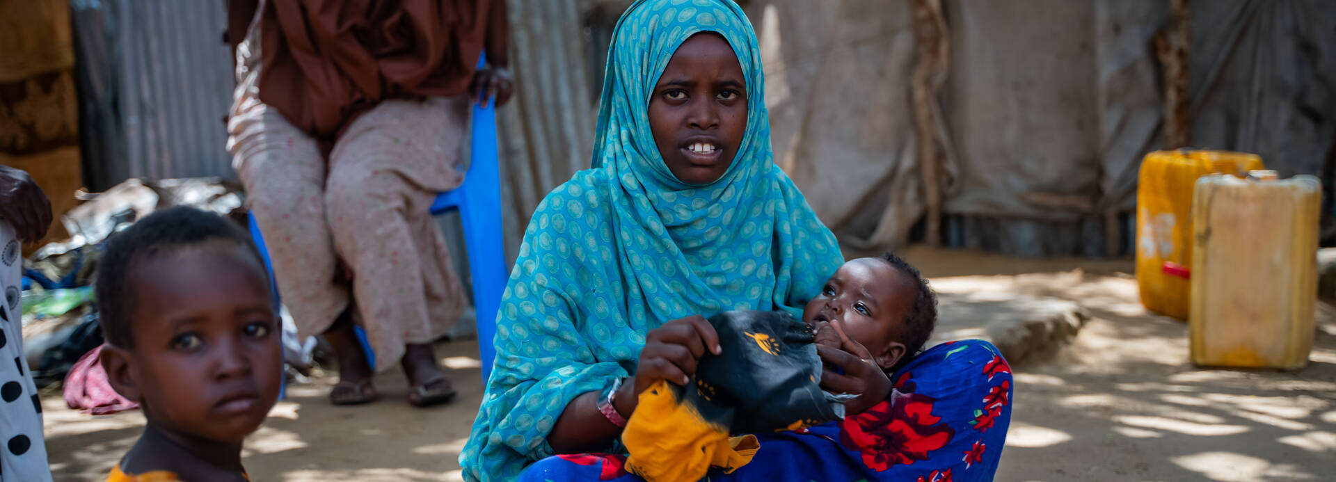 Eine junge somalische Mutter sitzt mit ihrem Baby auf dem Boden, im Vordergrund schaut ein Junge in die Kamera.
