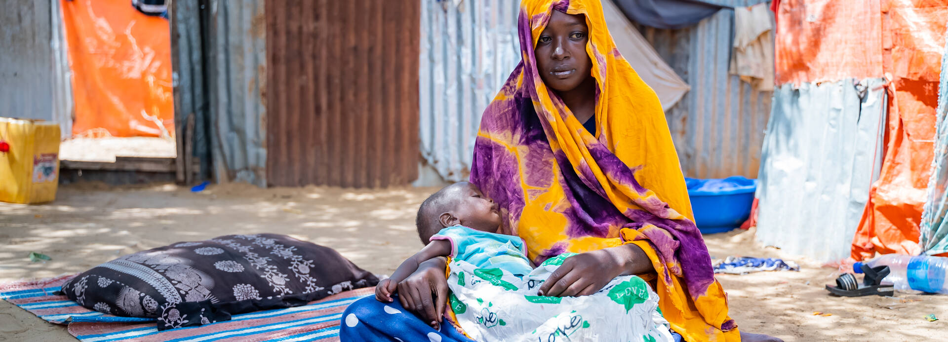 Eine junge Frau aus Somalia sitzt mit ihrem Baby in Tücher gehüllt auf einer Decke in einem Lager und schaut mit leerem Ausdruck zur Seite.