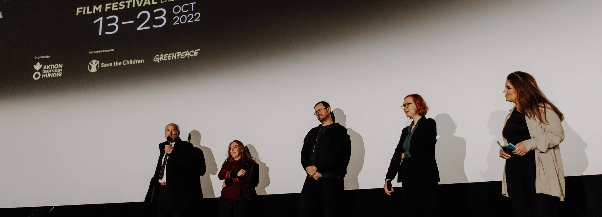 John Shipton, Stella Assange und Gabriel Shipton zusammen mit Anna Ramskogler-Witt und Moderatorin Sham Jaff auf der Bühne, auf der Leinwand das Logo des Human Rights Film Festival Berlin