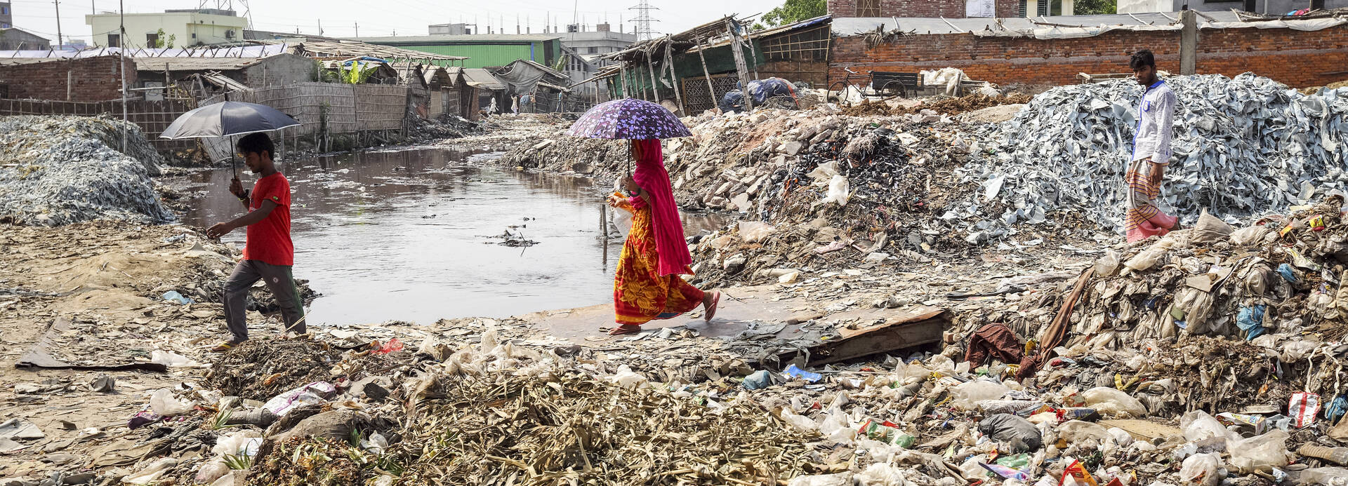 Drei Menschen aus Bangladesch laufen mit Schirmen durch ein Feld voller Abfälle einer Gerberei