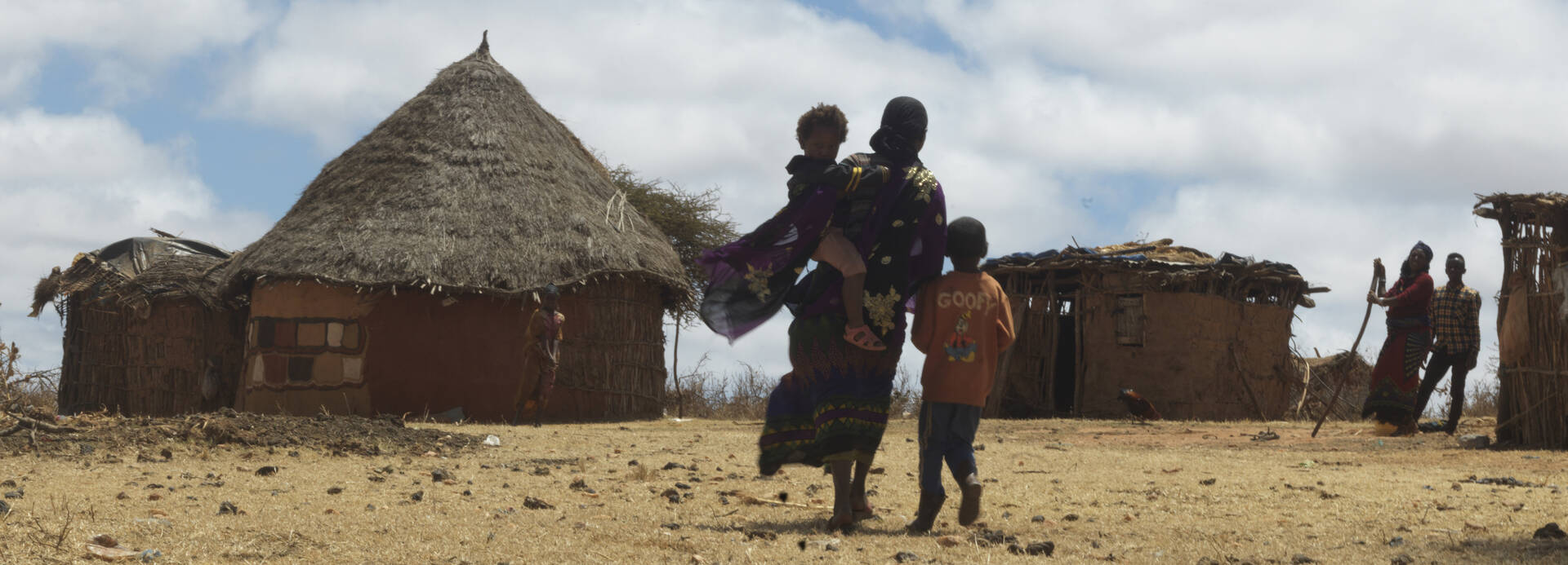 Eine Mutter läuft mit ihren Kindern durch die karge Landschaft Äthiopiens in ihr Dorf, im Hintergrund stehen zwei weitere Personen neben Hütten
