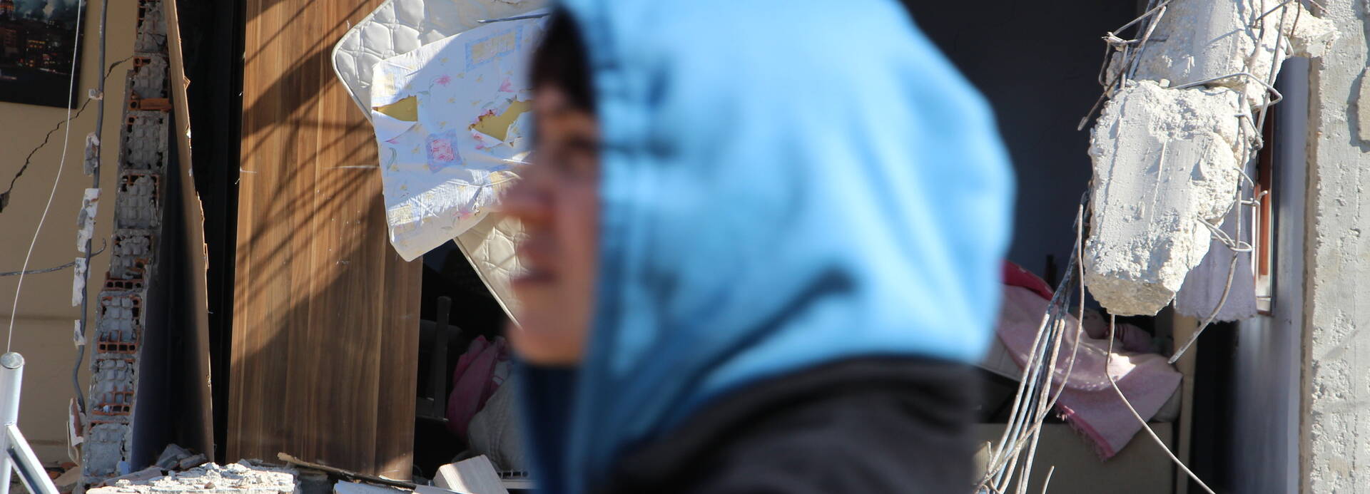 Das Ausmaß der Zerstörung nach den Erdbeben in der Türkei und in Syrien ist für die Menschen kaum fassbar. Vorne eine Person mit blauer Kapuze, unscharf, dahinter ein Teil eines zusammengestürzten Hauses, scharf